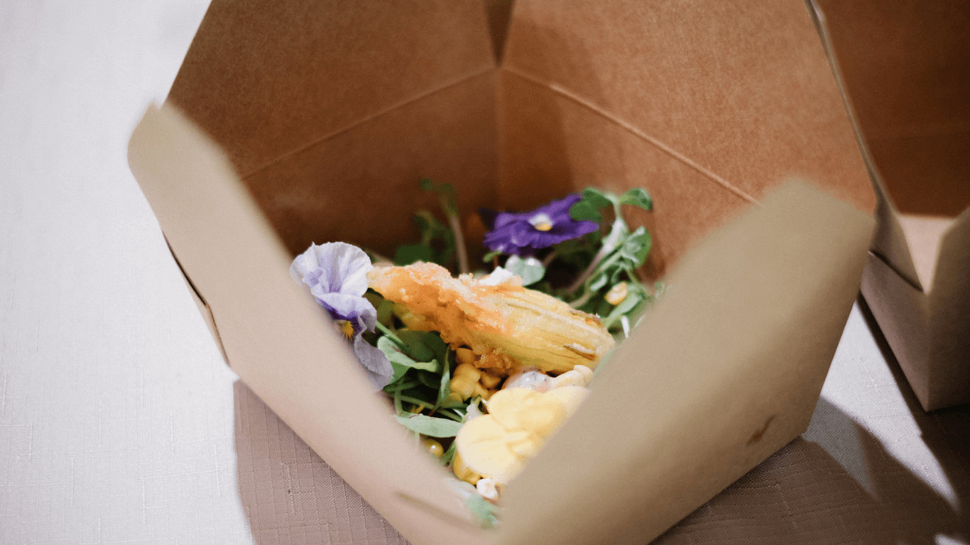 Eco-friendly takeout box with a fresh gourmet salad, garnished with edible flowers and a light fried zucchini blossom.